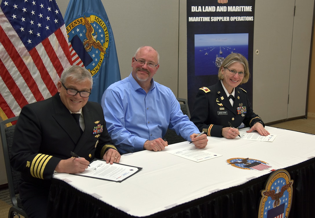 Three people sit at a table with a black skirt and the DLA logo. They are signing a contract like document. One is a woman in a dark Army uniform. She has short blond hair. The other is a man in a Army uniform. He has gray hair. Both are light skinned. There is a light skinned man in the middle with a blue dress shirt and gray hair and beard. All are smiling.