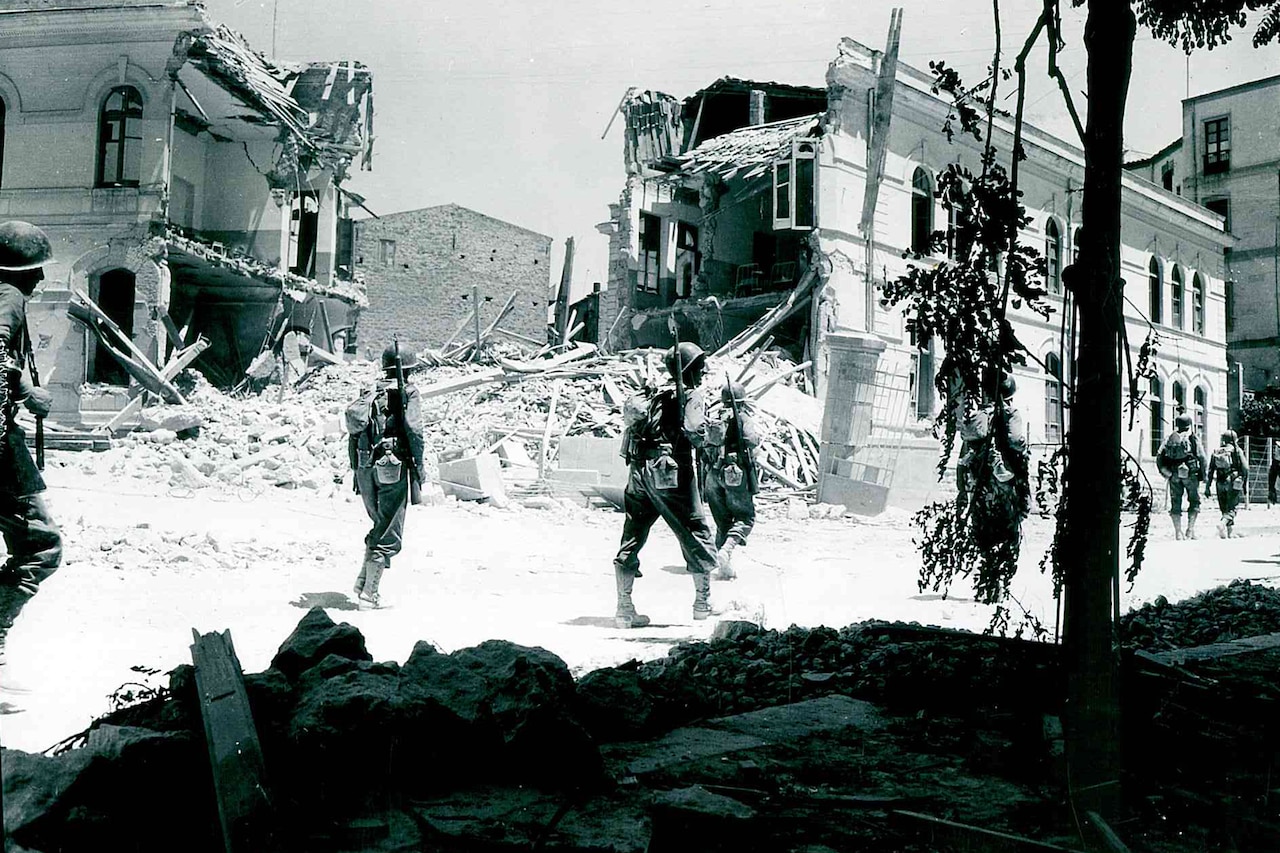 People in formation march past destroyed buildings in a town.