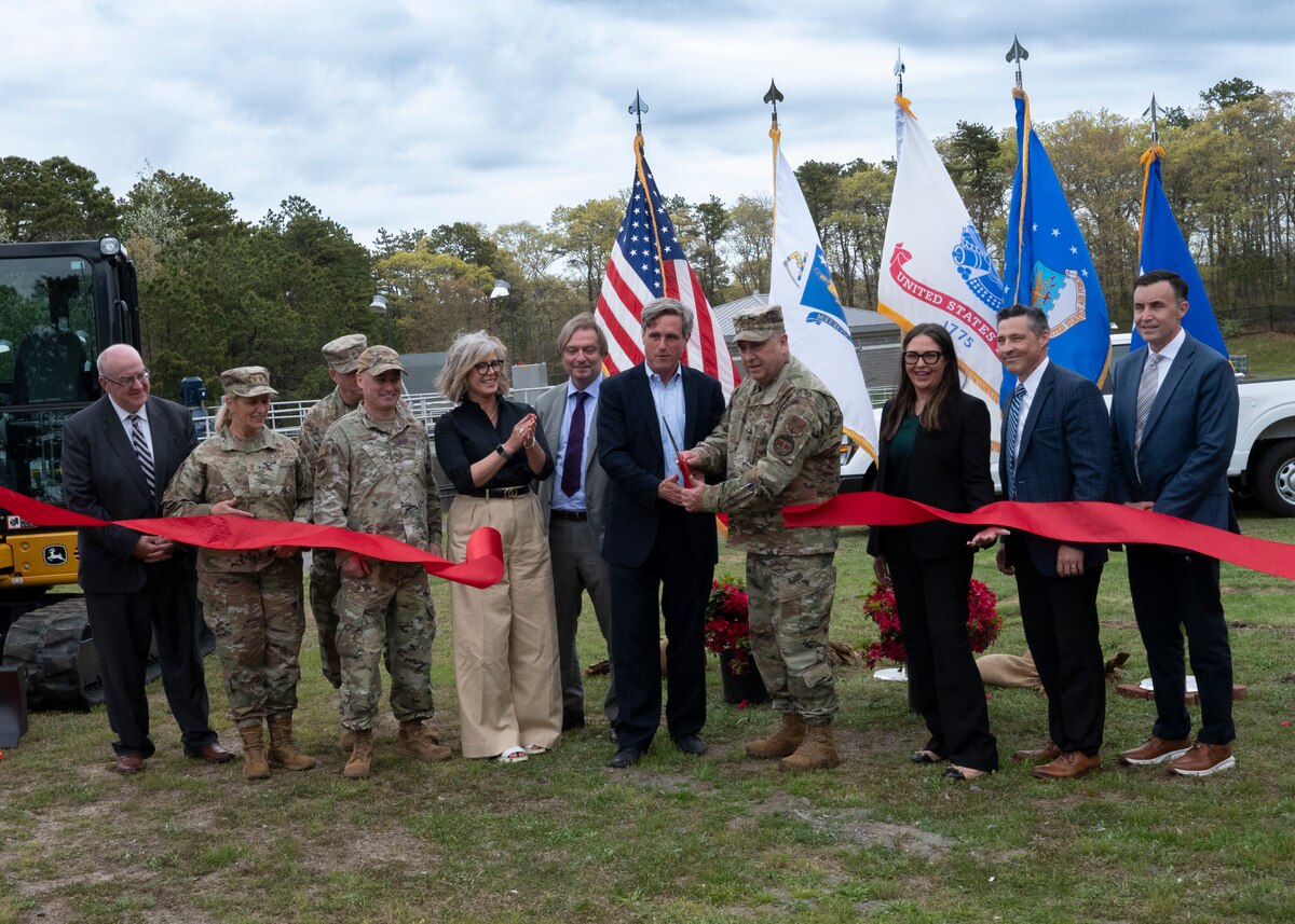 A group of military officers, community leaders and corporate management standing behind a large red ribbon as it is being cut