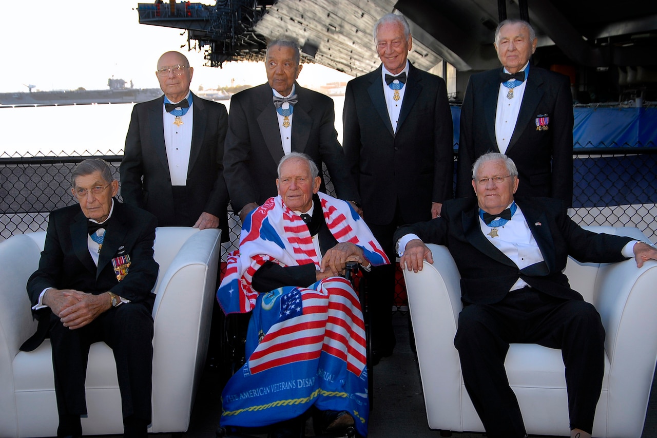 Seven people in tuxedos pose in two rows for a photo while on a ship.
