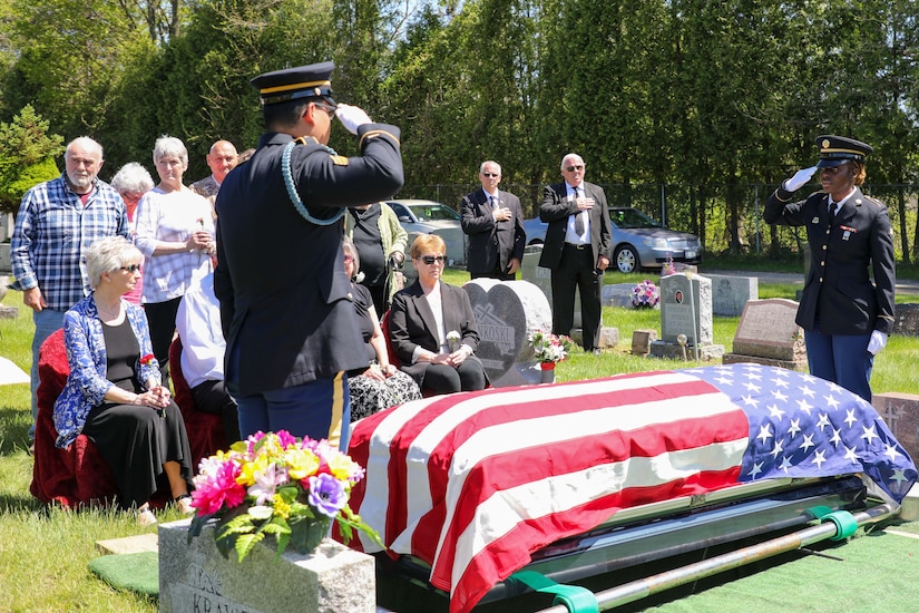 Soldiers salute  over a flag-draped casket while a group of people look on.