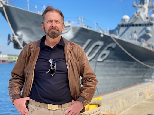 Man in blue/black shirt and brown jacket standing in front of ship with hands on hips.