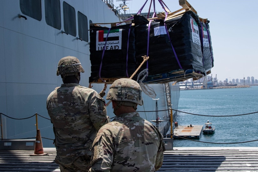 Two soldiers employ a guide rope on a container being lifted from a boat to a pier.