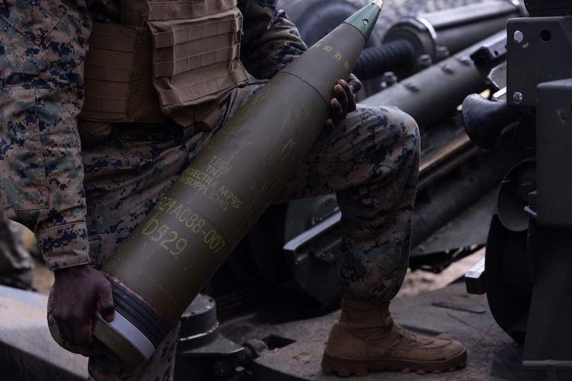 A service member in uniform holds an artillery round.