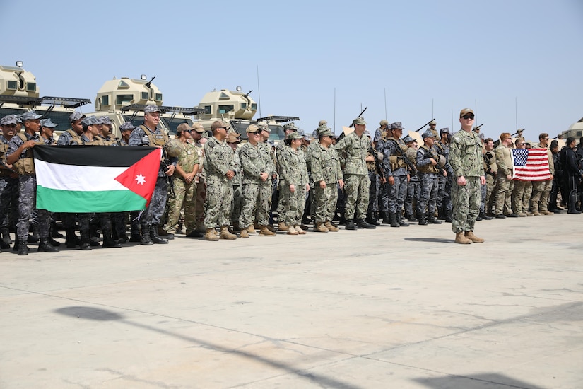 Dozens of service members in camouflage uniforms stand in a line in front of a trio of military vehicles. Some of the service members are holding up a black, white and green-striped flag; others hold a red and white-striped flag with 50 white stars over a blue background in the corner.