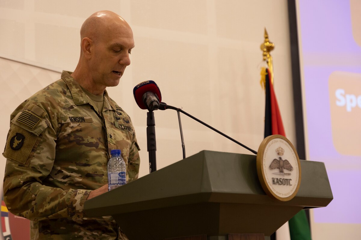 A man in a camouflage military uniform stands at a lectern with a microphone.