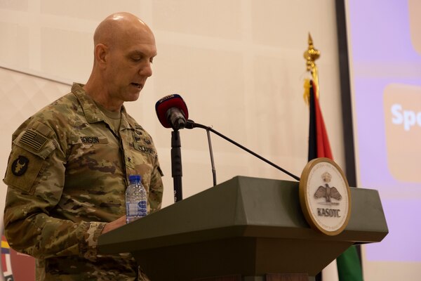 A man in a camouflage military uniform stands at a lectern with a microphone.