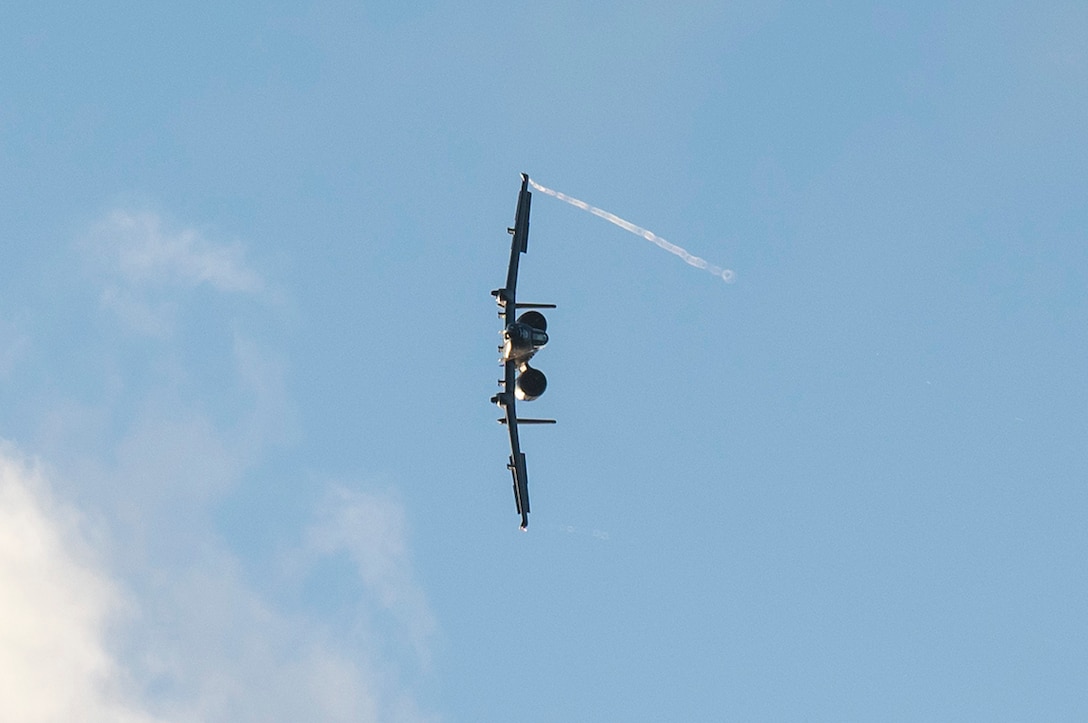 A military aircraft flies on its side during an air show, leaving a small trail of vapor.