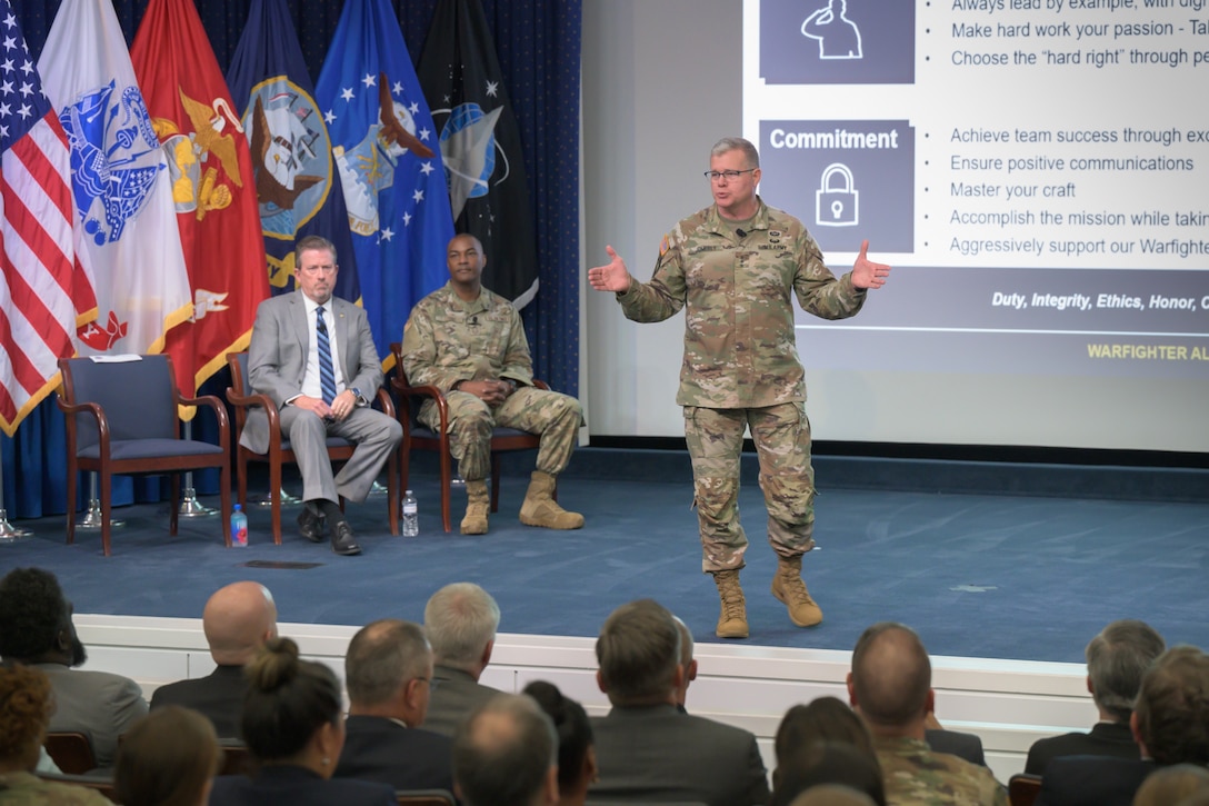 Army Lt. Gen Mark Simerly speaks from on stage with Brad Bunn and Chief Master Sgt. Alvin Dyer behind him. The slide in the background highlights the word "committment" behind Simerly