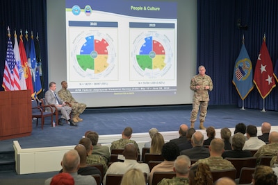 Lt. Gen Mark Simerly is on stage addressing a seated auditorium crowd with a comparison of two climate survey results displayed on a slide behind him