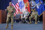 Lt. Gen Mark Simerly stands on stage with Brad Bunn and Chief Master Sgt. Alvin Dyer seated behind him with a background of flags