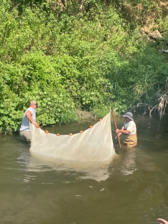 Two men stand in waist high water with a net in between them and trees in the background.