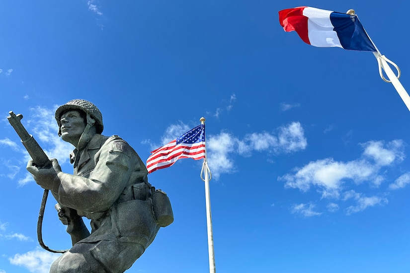 A statue of a uniformed service member holding a weapon stands between a U.S. flag and a French flag.