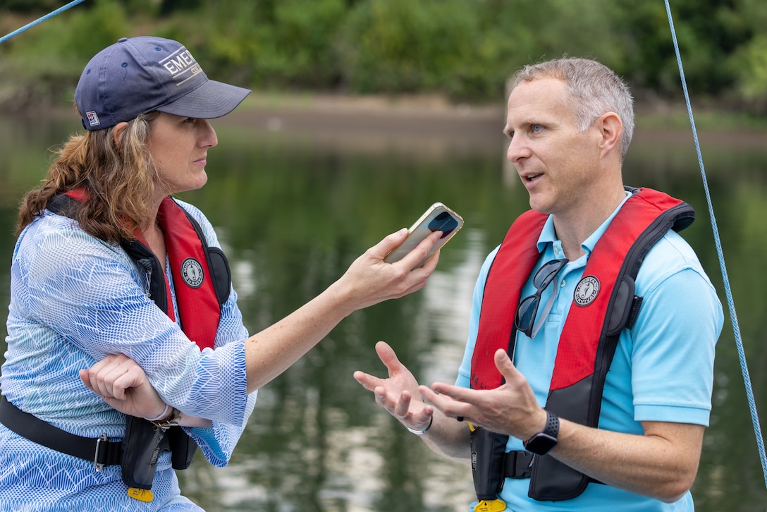 Woman on the left-side of picture holds phone to record conversation with man on right-side of image.