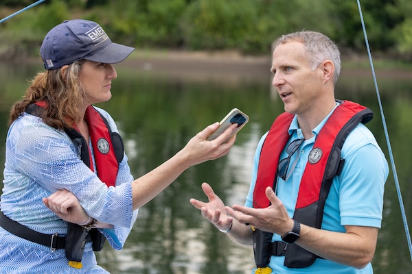 Woman on the left-side of picture holds phone to record conversation with man on right-side of image.