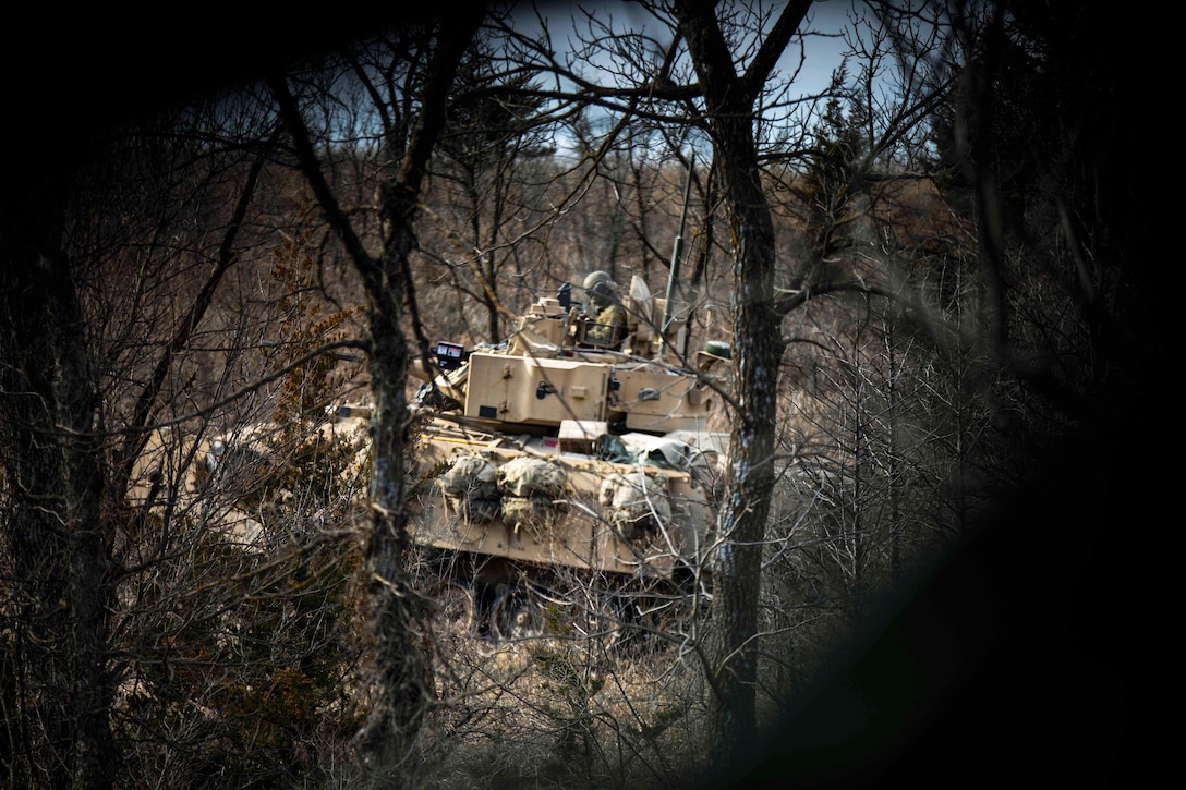 A soldier operates a tank in the forest as seen through branches.