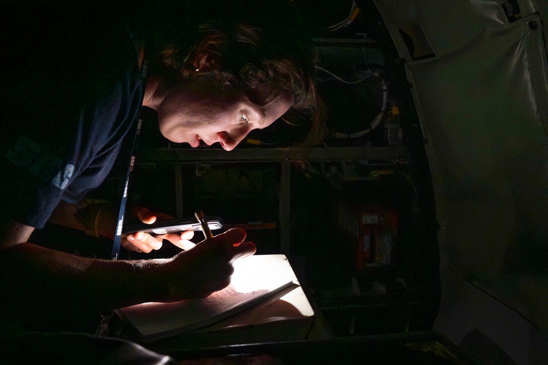 An airman bends over while using their phone's flashlight to write in a journal in the dark on an aircraft.