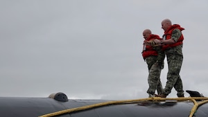 Two service members wearing life vests walk on the roof of a submarine.