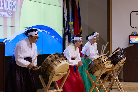 Four women in white shirts and headbands play drums.
