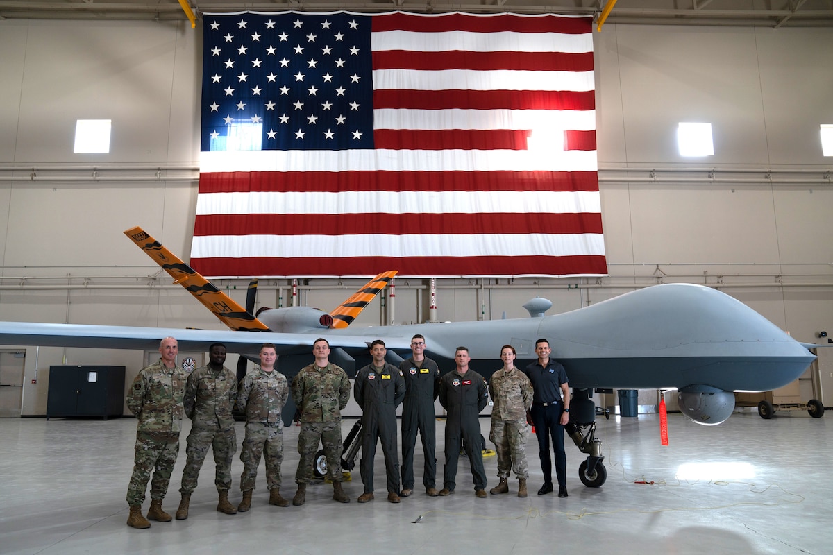 A group of military members in uniform pose for the camera in front of a remotely piloted aircraft.
