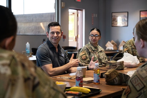 A man in a polo shirt sitting at a table talks to military members in uniform.