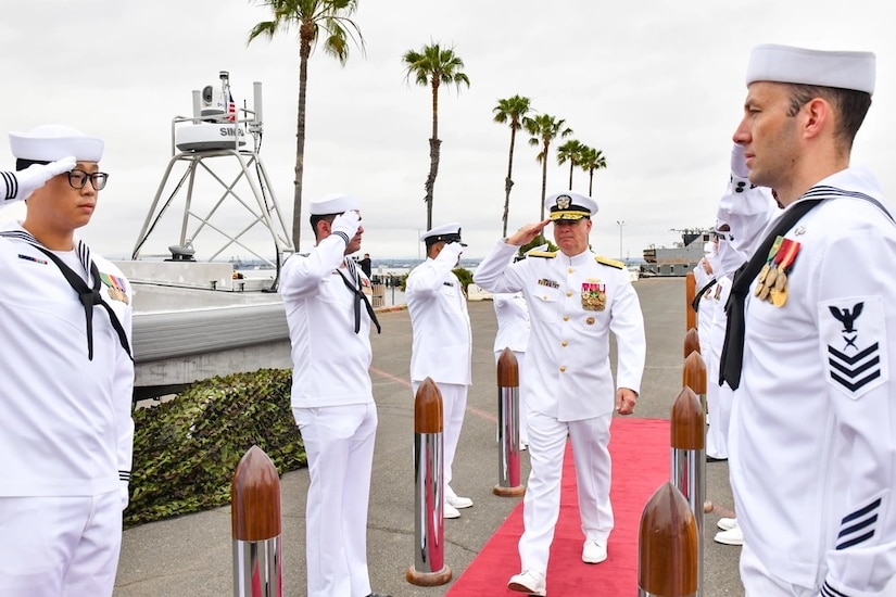 A sailor in dress whites walking on a red carpet outdoors salutes fellow sailors standing in two rows.