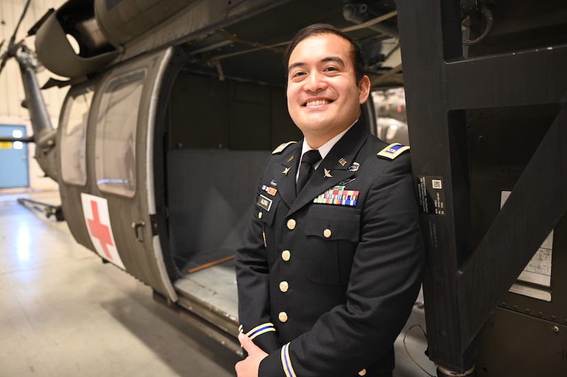 An Army warrant officer stands and smiles in front of a parked Black Hawk helicopter inside a building.