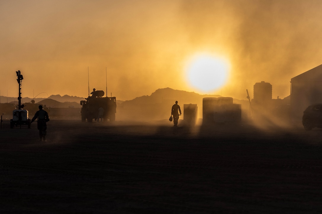 Service members and vehicles move through a sandy, mountainous region as the sun shines through the dusty air creating a golden glow.