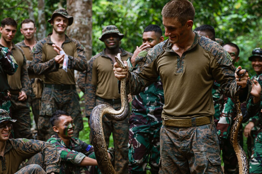 A Marine holds a snake in a jungle setting while other service members smile and watch.