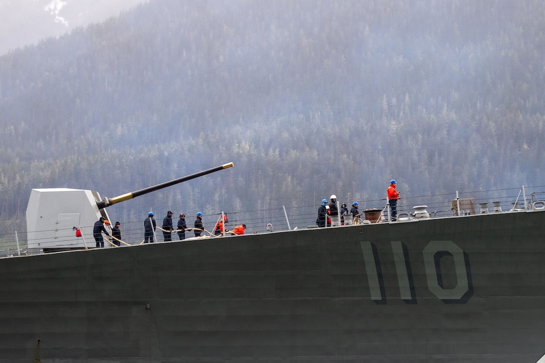 A group of sailors hold mooring line on the deck of a Navy ship on an overcast day.