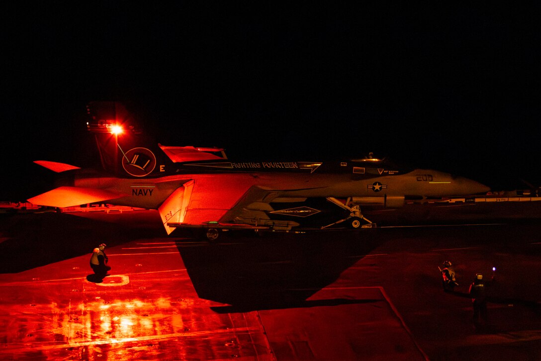 Sailors signal a military aircraft on the flight deck of a Navy ship at night. The deck is illuminated by an orange light.