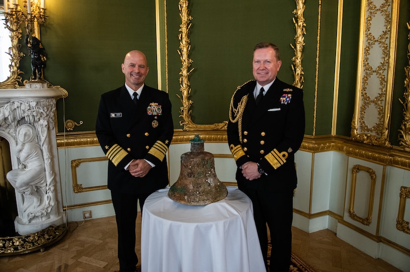 Two uniformed service members stand next to a World War I ship's bell.
