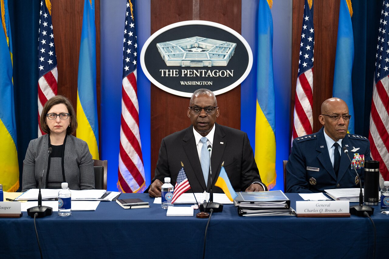 Secretary of Defense Lloyd J. Austin III sits at a table between Air Force Gen. CQ Brown, Jr., and a civilian in front of U.S. and Ukrainian flags.