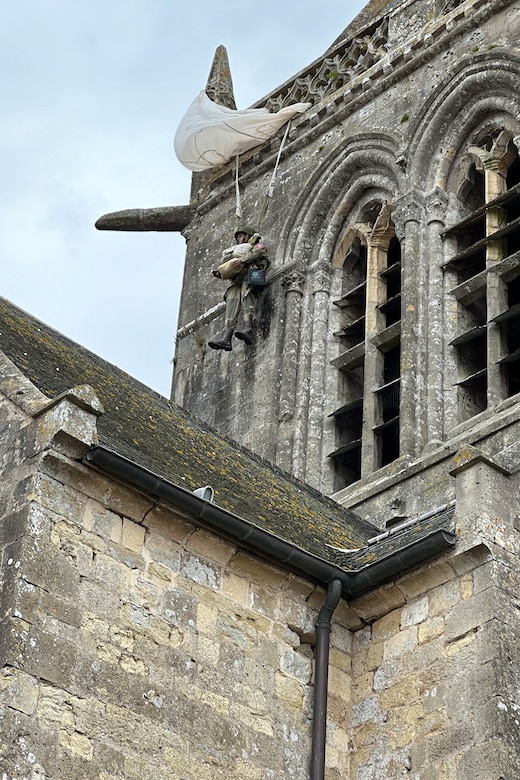 A mannequin with parachute hangs from a church steeple.