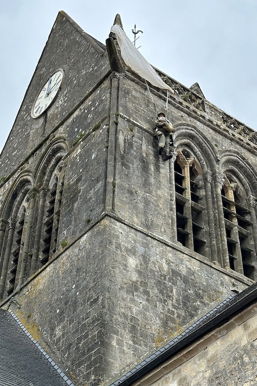 Mannequin with parachute hangs from church steeple.