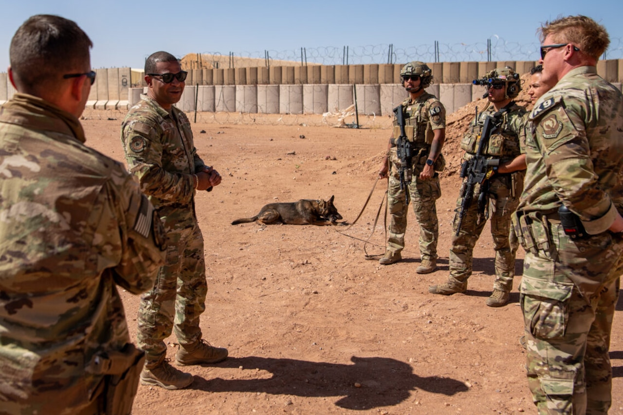 A man in uniform talks to surrounding men and woman in uniform. A dog on a leash lays down on the dirt next to his handler.