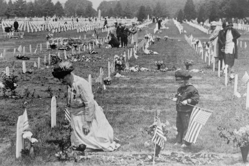 A person decorates a grave while a child holding an American flag watches.