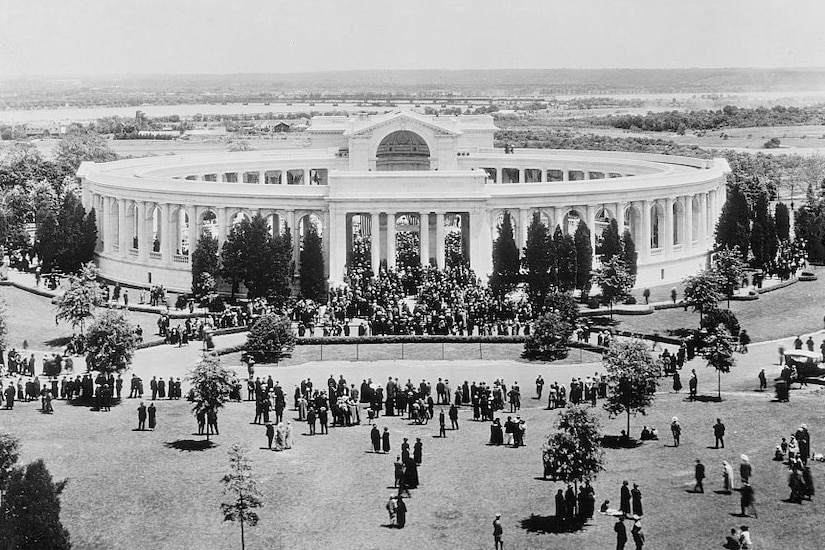 A large round building is seen with people standing around it.