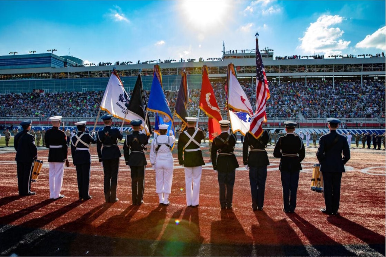 A military color guard stands on a track facing a massive set of stands filled with people.