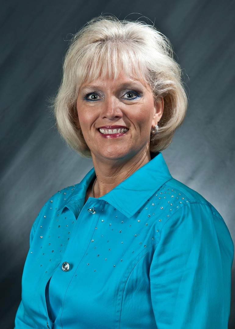 A light-skinned woman with white hair wearing a blue dress shirt is smiling at the camera.