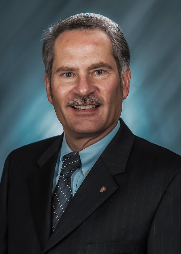 A light-skinned man with greying hair wearing a dark suit is smiling at the camera.