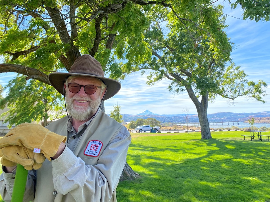 Man in a wide brim hat, stands in front of green grass and beautiful trees.