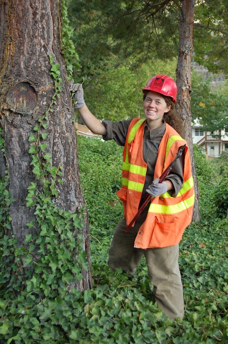 Women in a red SCA hard hat and an orange and yellow safety vest stands in front of a tree.