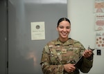 Uniformed service member smiles and holds clipboard in front of door that says "Mental Health."