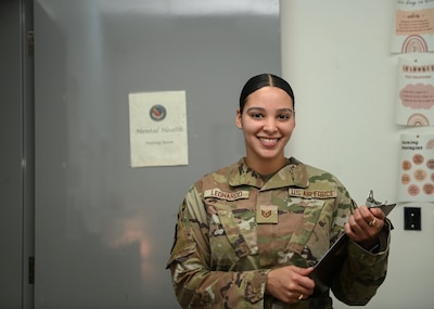 Uniformed service member smiles and holds clipboard in front of door that says "Mental Health."