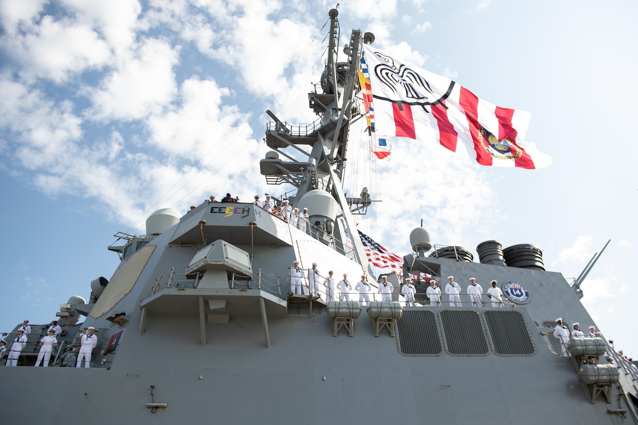 Sailors in formal uniforms stand at attention along the rails of a large ship as seen from below. A large flag flies above them.