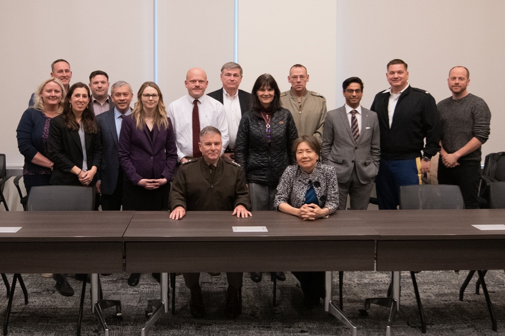 Lieutenant General Hartman sits at a table with DARPA director Stefanie Tompkins while members of the program gather behind them for a group photo.