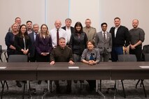 Lieutenant General Hartman sits at a table with DARPA director Stefanie Tompkins while members of the program gather behind them for a group photo.