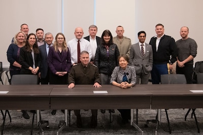 Lieutenant General Hartman sits at a table with DARPA director Stefanie Tompkins while members of the program gather behind for a group photo.