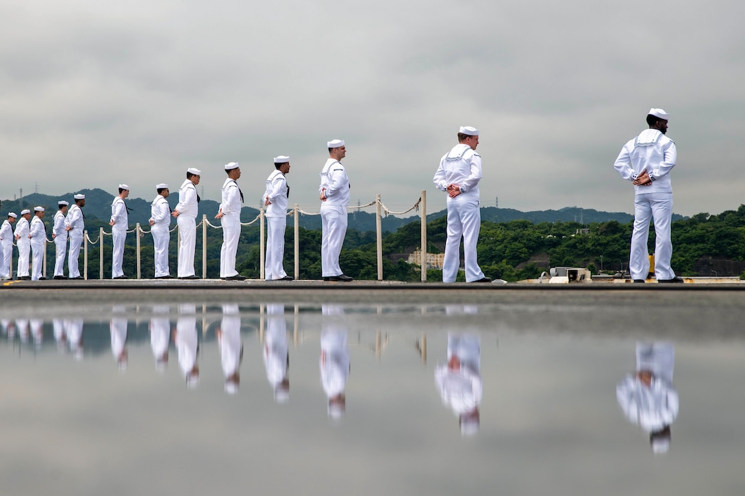 A dozen sailors in uniform stand in formation on the edge of a ship facing mountains as their reflections are seen in a puddle on the flight deck.